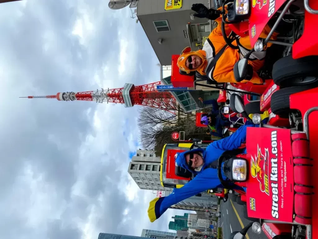 People in colorful costumes ride red go-karts on a city street, with a tall red-and-white tower visible in the background.