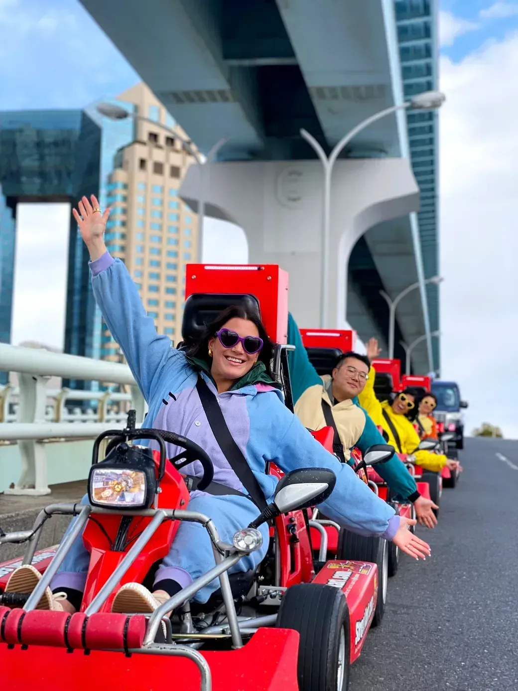 Smiling woman in a blue jacket and purple sunglasses waves from a red go-kart on a city street, with others in line behind under a large bridge.