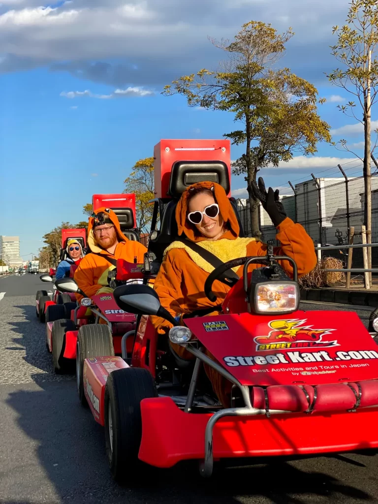 Front rider in an orange mascot suit with white heart-shaped sunglasses waves from a red go-kart in a street parade; blue sky and trees in background.