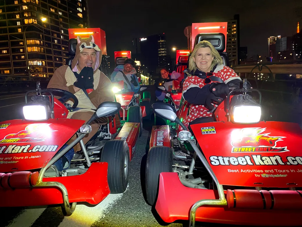 Nighttime go-kart photo with two costumed drivers in front on a city street, red karts and bright headlights.